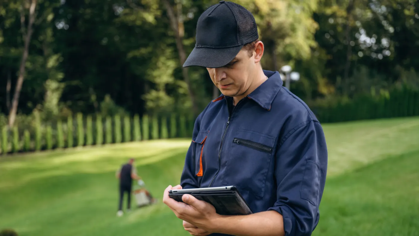 A landscaping professional in a blue uniform and black cap stands on a large, green lawn and looks at a tablet, while a coworker mows the grass in the background.