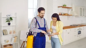 A professional pest control technician discusses the service quote on a clipboard with a female homeowner in her kitchen, before starting the fumigation treatment.