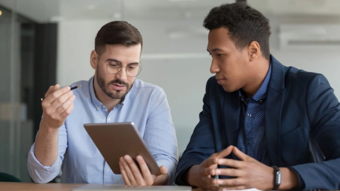 two men sitting at a desk, discussing while one holds a tablet and the other looks at it