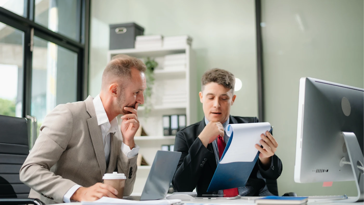 two people sitting at a desk, discussing and reviewing a paper or report together.
