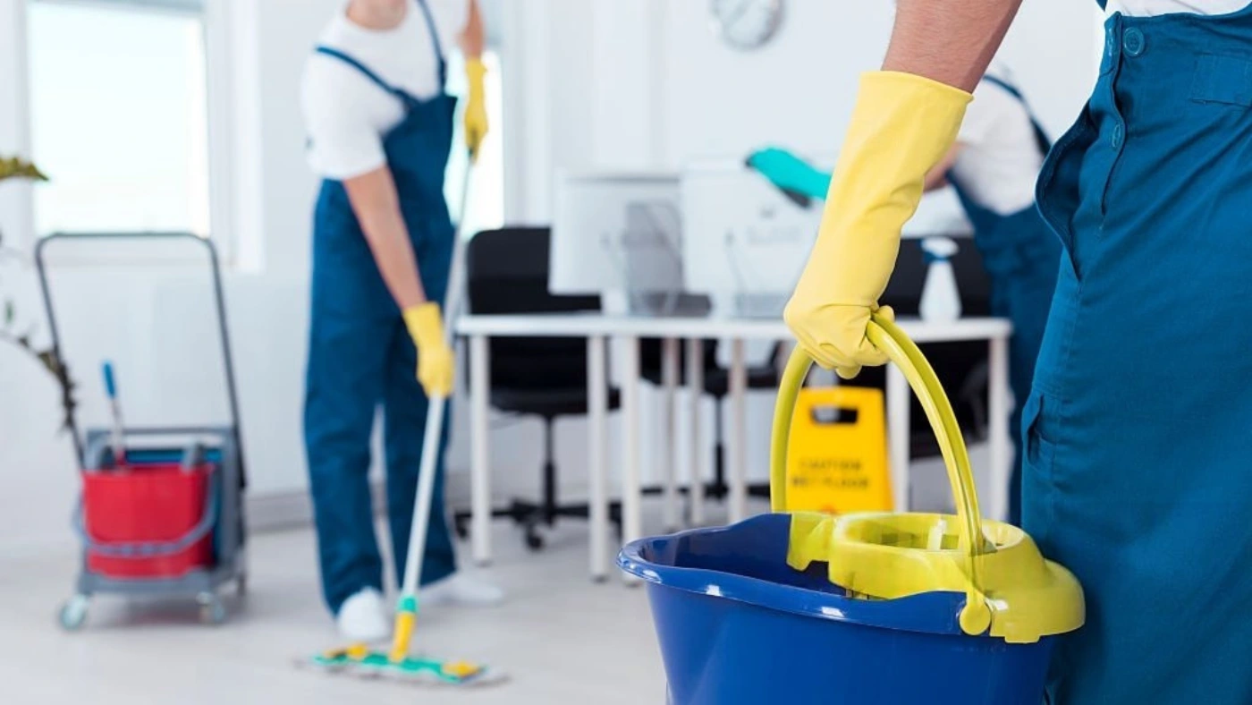 two cleaning professionals are tidying an office; one is mopping the floor while the other wipes a desk, and a cleaner holds a mop bucket nearby