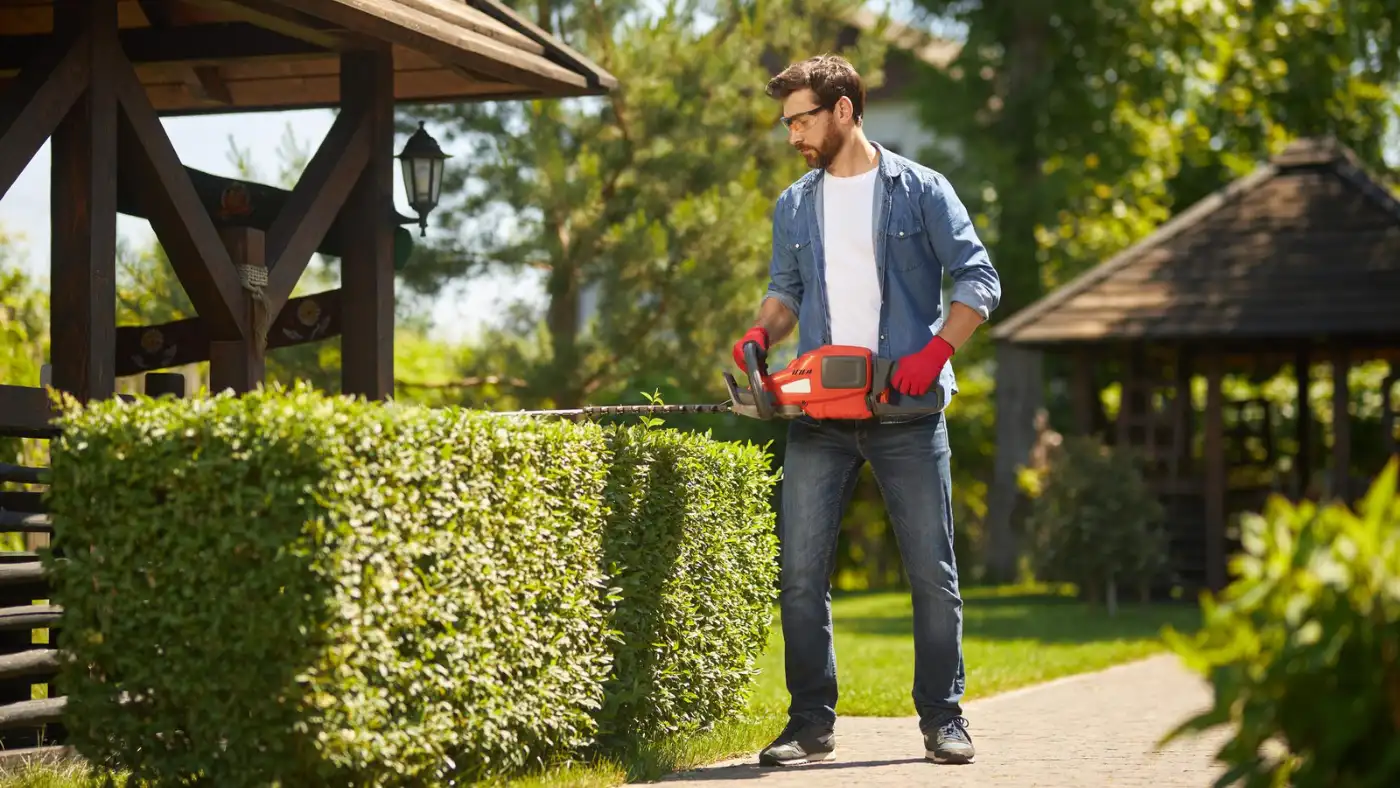 Professional landscaper trimming hedges with a power hedge trimmer in a residential yard.